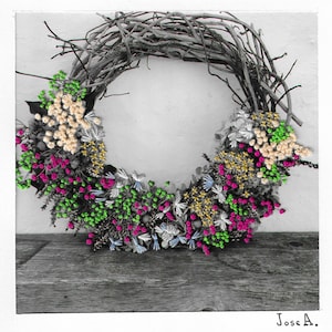 Op de afbeelding: Een handgemaakte krans met een basis van natuurlijke twijgen, versierd met kleurrijke bloemelementen. De krans toont clusters van roze, crème, gele, groene en blauwe bloemen, waardoor een levendig en gestructureerd ontwerp ontstaat. De krans wordt gepresenteerd tegen een neutrale achtergrond.