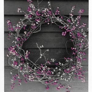 Op de afbeelding: Een cirkelvormige krans gemaakt van verweven takken, versierd met kleine roze en witte bessen en bloemen. De krans staat tegen een donkergrijze houten achtergrond. De handtekening van de kunstenaar, "Jose A.", is rechtsonder zichtbaar.