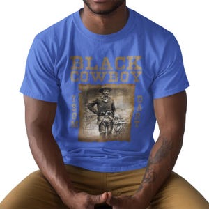 May include: Royal blue t-shirt featuring a sepia-toned image of a Black cowboy. The text "BLACK COWBOY" is printed above the image in a gold color, with additional text on the sides. The shirt is made of a soft material.