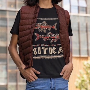 May include: A black t-shirt with a graphic of two stylized salmon fish and the word "SITKA" in a decorative font. The shirt is being worn by a person with long brown hair and a brown vest.