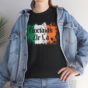 以下が含まれることがあります： 白地に「Tiocfaidh ár lá」の文字が入った、くたびれたアイルランド国旗のデザインが施された黒いTシャツです。