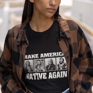 May include: A black t-shirt with a white graphic that says "Make America Native Again". The graphic features four black and white images of Native American people. The person wearing the shirt is standing in front of a brown and black checkered background.