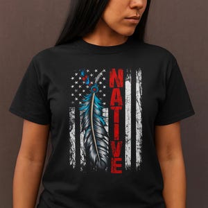 Könnte beinhalten: Schwarzes T-Shirt mit einem indianischen Federdesign. Die Feder ist blau, grau und rot, vor einem Hintergrund der zerrissenen amerikanischen Flagge. Das Wort "NATIVE" ist vertikal in Rot gedruckt.