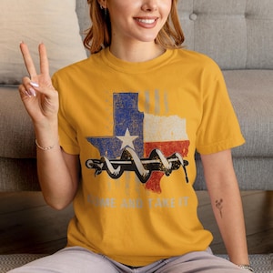 Könnte beinhalten: Senfgelbes T-Shirt mit einem verwitterten Texas-Flaggen-Design, einer zusammengerollten Schlange und dem silbernen Text "COME AND TAKE IT". Die Texas-Flagge ist blau, weiß und rot. Das T-Shirt hat kurze Ärmel.