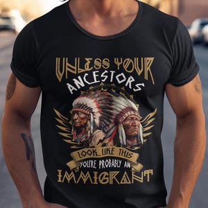 Könnte beinhalten: Schwarzes T-Shirt mit Rundhalsausschnitt und einer Grafik von zwei indianischen Figuren mit Kopfschmuck. Der Text lautet "UNLESS YOUR ANCESTORS LOOK LIKE THIS YOU'RE PROBABLY AN IMMIGRANT" in goldfarbenen Buchstaben.