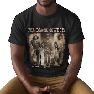 May include: Black t-shirt featuring a sepia-toned graphic of three cowboys with the text "THE BLACK COWBOYS" above them. The names "NAT LOVE", "ISOM DART", and "BILL PICKETT" are printed below the cowboys. The shirt is made of a soft material.