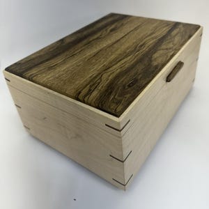 Op de afbeelding: Een rechthoekige houten kist met een lichtgekleurde body en een donker, patroonvormig deksel. De kist heeft zwaluwstaartverbindingen op de hoeken en een kleine, donkere houten handgreep. Het deksel heeft een rijke houtnerf.