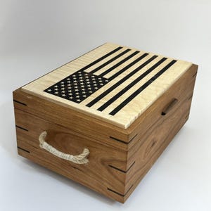 Op de afbeelding: Een houten kist met een deksel met een ontwerp van de Amerikaanse vlag. Het deksel is lichtgekleurd hout met zwarte strepen en sterren. De kist is gemaakt van donkerder hout met een touwhandvat aan de zijkant.