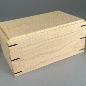 May include: A rectangular wooden box crafted from light-colored curly maple wood. The lid features a distinct wavy grain pattern. The corners of the box are joined with dark wood splines, providing a visible contrast against the pale timber.