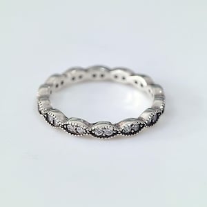 Könnte beinhalten: Ein silberner Ring mit einem sich wiederholenden Muster aus ovalen Fassungen, die jeweils kleine, klare Edelsteine enthalten. Der Ring hat ein zartes, verziertes Design mit poliertem Finish. Die Edelsteine funkeln auf dem silbernen Band.