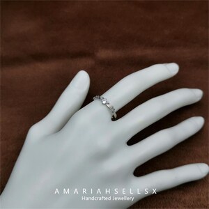 May include: A silver ring with a delicate, wavy design and small clear stones. The ring is on a hand, with the text "AMARIAHSELLSX Handcrafted Jewellery" below.