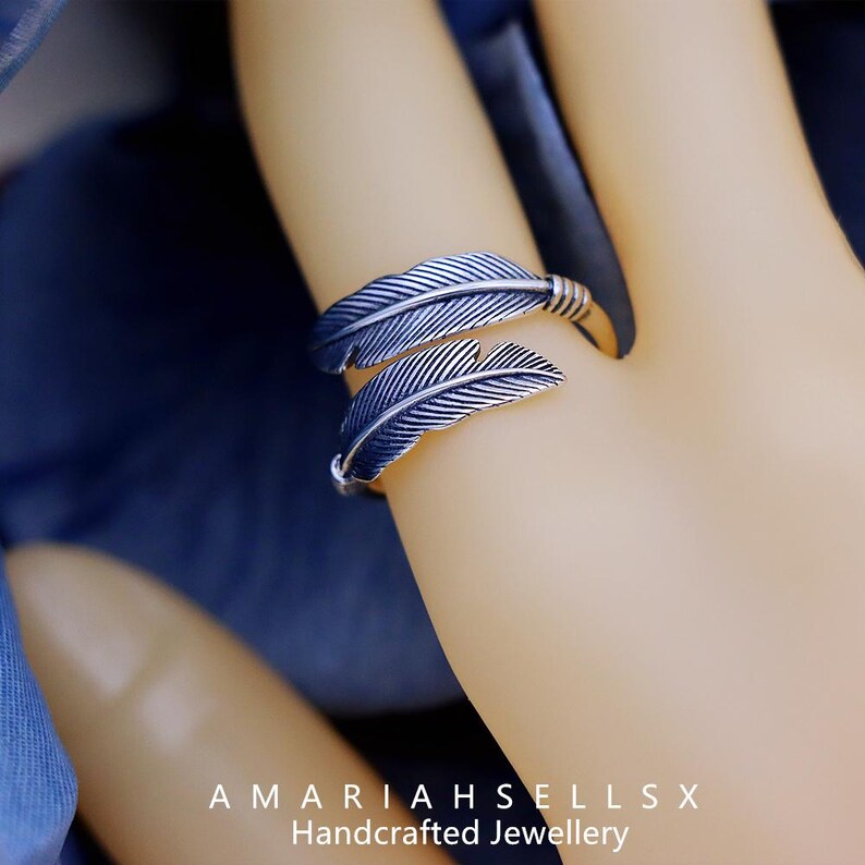 Puede incluir: Un anillo plateado con dise&ntilde;o de pluma. El anillo tiene dos formas de plumas superpuestas con vetas detalladas. La banda es ajustable y est&aacute; envuelta con metal texturizado. La imagen incluye el texto "AMARIAHSELLSX Handcrafted Jewellery".