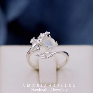 以下が含まれることがあります： 白い楕円形の宝石と繊細な花のデザインが施されたシルバーリング。リングは手作りで、小さな花が咲いた枝が特徴です。