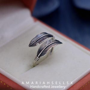 Puede incluir: Anillo plateado con dise&ntilde;o de plumas. El anillo tiene dos plumas detalladas que envuelven el dedo. El anillo se muestra en una caja de joyer&iacute;a roja y blanca. El texto "AMARIAHSELLSX Handcrafted Jewellery" est&aacute; en la parte inferior.