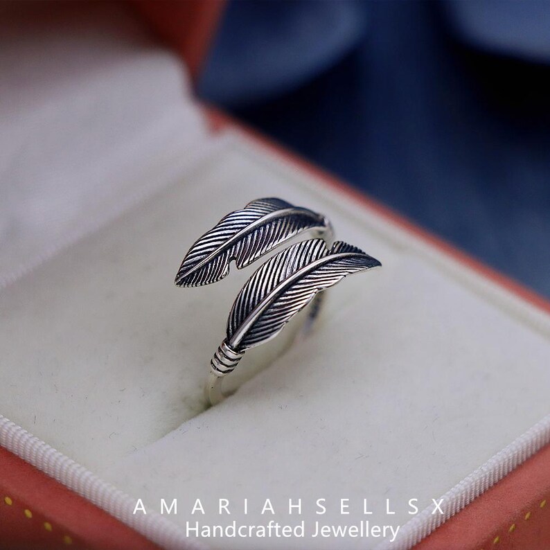 Puede incluir: Anillo plateado con dise&ntilde;o de plumas. El anillo tiene dos plumas detalladas que envuelven el dedo. El anillo se muestra en una caja de joyer&iacute;a roja y blanca. El texto "AMARIAHSELLSX Handcrafted Jewellery" est&aacute; en la parte inferior.