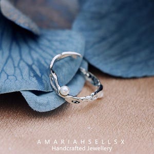 May include: A silver ring with a textured, organic design and a small white pearl accent. The ring is on a blue background.