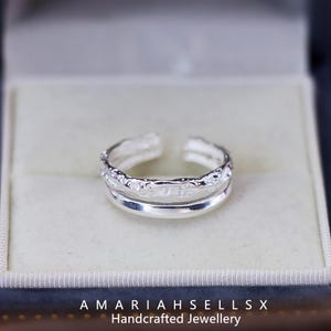 May include: A silver adjustable ring with a textured band and a smooth band layered on top. The ring is in a white box with the text "AMARIAHSELLSX Handcrafted Jewellery".