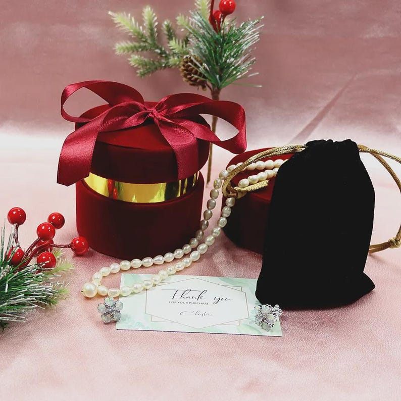 Puede incluir: Una caja de regalo de terciopelo rojo con una banda dorada y un lazo de cinta de raso. Se exhiben un collar y pendientes de perlas con una bolsa de terciopelo negro y una tarjeta que dice "Thank you". Decoraci&oacute;n navide&ntilde;a decorativa en el fondo.