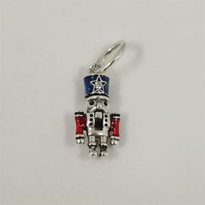 May include: A silver-colored charm in the shape of a nutcracker soldier. The charm has a blue hat with a star, red arms, and a detailed face. The charm is designed to be attached to a bracelet or necklace.
