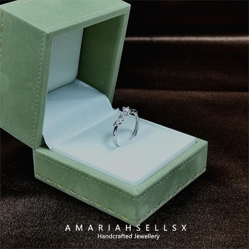 May include: A silver ring with a delicate design and small clear stones, displayed in a green velvet ring box. The box has the text "AMARIAHSELLS" and "Handcrafted Jewellery" on the bottom.