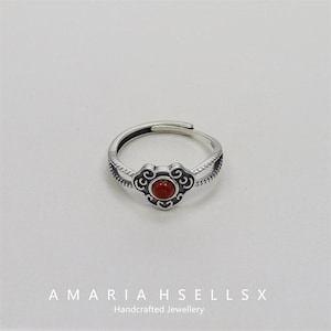 Pode incluir: Anel prateado com um design central em forma de coração com uma pequena pedra vermelha redonda. O anel é ajustável e a banda tem um padrão texturizado. O texto "AMARIA HSELLSX Handcrafted Jewellery" está na parte inferior.