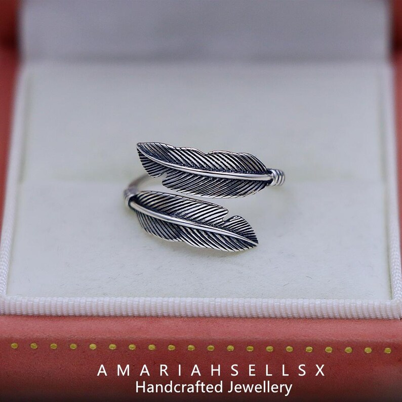 Puede incluir: Anillo plateado con dise&ntilde;o de plumas, presentado en una caja de joyer&iacute;a rosa. El anillo presenta dos plumas detalladas. El dise&ntilde;o del anillo envuelve el dedo. El texto "AMARIAHSELLSX Handcrafted Jewellery" es visible.