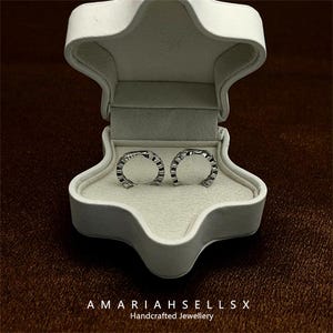 May include: A pair of silver hoop earrings with a geometric design, displayed inside a white jewelry box. The box is open, revealing the earrings against a cream-colored interior. The text "AMARIAHSELLSX Handcrafted Jewellery" is at the bottom.