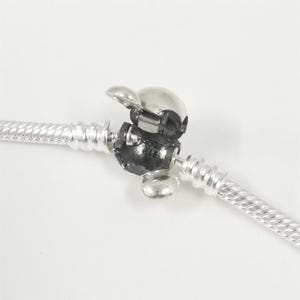 Puede incluir: Un cierre de pulsera plateado con una parte superior redondeada con bisagras y una base negra. El cierre est&aacute; unido a una pulsera plateada trenzada estilo cuerda. El fondo es blanco liso.