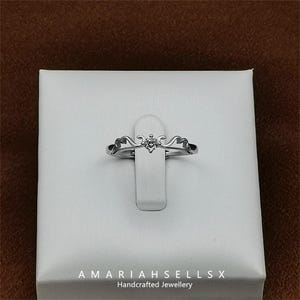 May include: A silver ring with a delicate, ornate design and a single, small, clear gemstone. The ring is displayed on a white background with the text "AMARIAHSELLSX Handcrafted Jewellery".