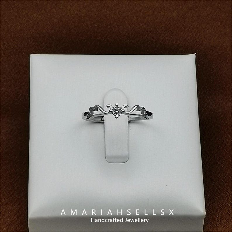 May include: A silver ring with a delicate, ornate design and a single, small, clear gemstone. The ring is displayed on a white background with the text "AMARIAHSELLSX Handcrafted Jewellery".
