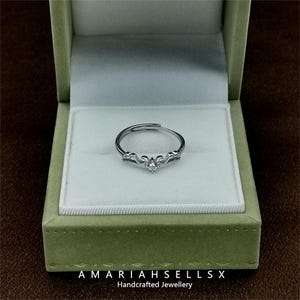 May include: A silver ring with a delicate design and a single clear stone in the centre. The ring is displayed in a green velvet ring box with the text "AMARIAHSELLS" and "Handcrafted Jewellery".