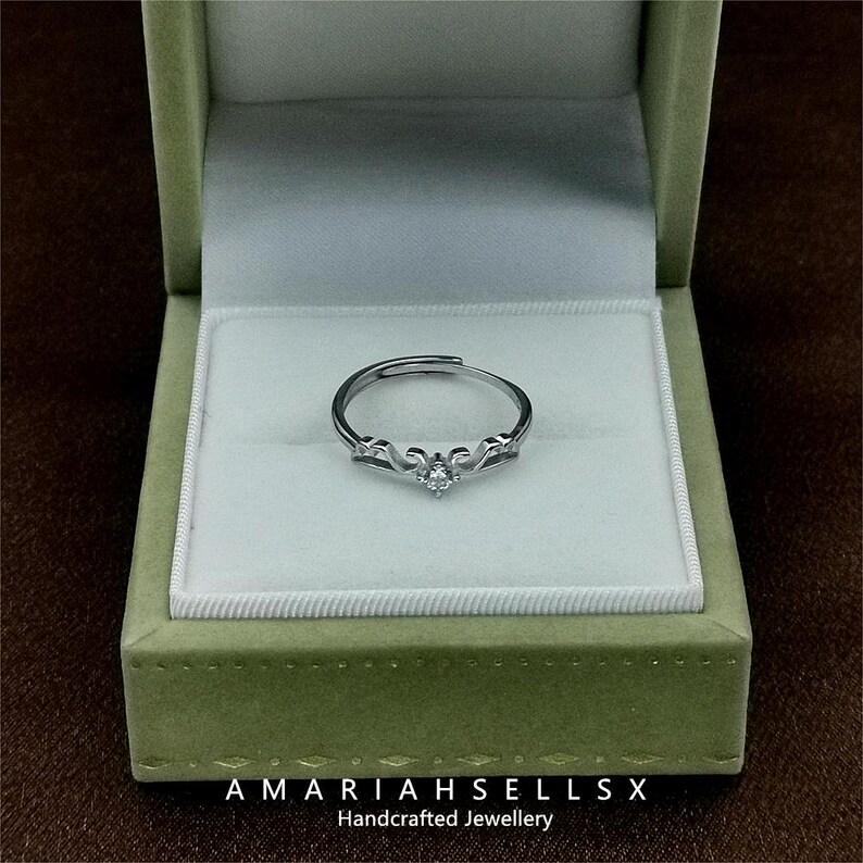 May include: A silver ring with a delicate design and a single clear stone in the centre. The ring is displayed in a green velvet ring box with the text "AMARIAHSELLS" and "Handcrafted Jewellery".