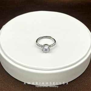 May include: A silver ring featuring a large, clear, round cubic zirconia stone surrounded by smaller cubic zirconia stones.