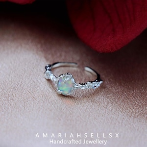 May include: A silver ring with a textured band and a round opal gemstone. The opal is iridescent with shades of blue, green, and pink.