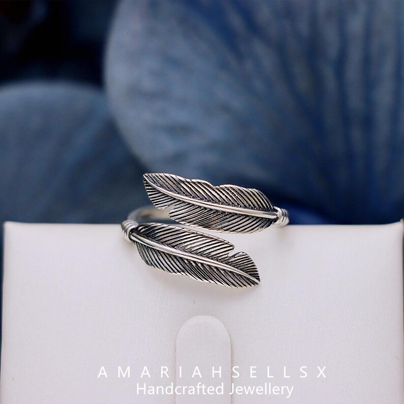 Puede incluir: Anillo plateado con dise&ntilde;o de plumas. El anillo presenta dos detalles de plumas. Se muestra sobre una superficie blanca con el texto "AMARIAHSELLSX Handcrafted Jewellery".