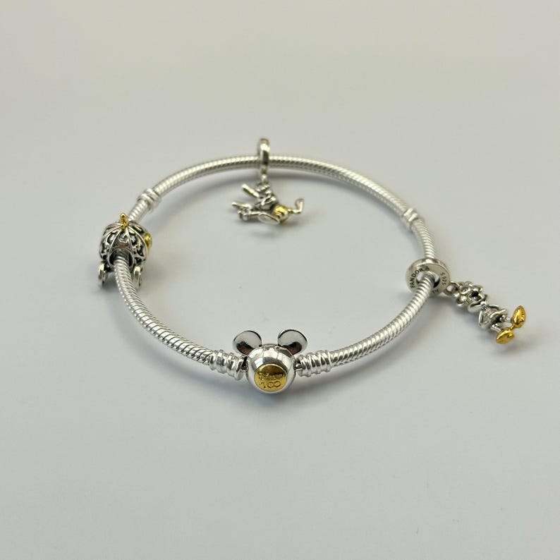 K&ouml;nnte beinhalten: Ein silbernes Armband mit mehreren Anh&auml;ngern. Zu den Anh&auml;ngern geh&ouml;ren eine Krone, ein Mickey Mouse-Kopf und eine Mickey Mouse-Figur mit goldenen Akzenten. Das Armband liegt auf einer wei&szlig;en Oberfl&auml;che.