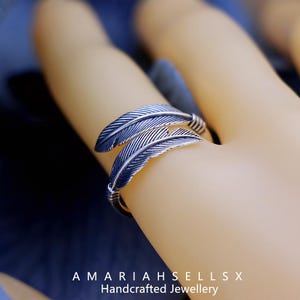 Puede incluir: Un anillo plateado con dise&ntilde;o de pluma. El anillo presenta dos formas de pluma detalladas que se envuelven alrededor del dedo. El anillo est&aacute; hecho a mano y el texto "AMARIAHSELLSX Handcrafted Jewellery" es visible.