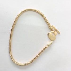 May include: A gold bracelet with a snake chain design. The bracelet has a heart-shaped charm and a T-bar clasp. The charm is engraved with the word "Pandora".