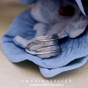 Puede incluir: Un anillo plateado con dise&ntilde;o de pluma con dos dise&ntilde;os de plumas detallados. El anillo est&aacute; sobre un fondo azul. El texto "AMARIAHSELLSX Handcrafted Jewellery" es visible en la parte inferior.