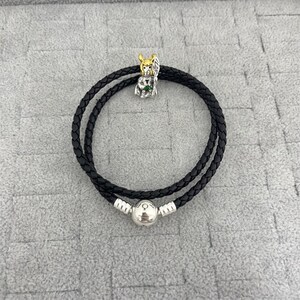 Könnte beinhalten: Schwarzes Lederarmband mit Silberverschluss und einem Charm mit einer goldfarbenen Figur mit grünen Akzenten. Das Armband ist doppelt gefädelt und hat eine geflochtene Textur.