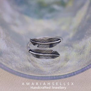 Puede incluir: Un anillo plateado con dise&ntilde;o de pluma. El anillo presenta dos motivos de plumas detallados, uno arriba y otro abajo, con l&iacute;neas intrincadas. El anillo se muestra sobre una superficie reflectante. El texto "AMARIAHSELLSX Handcrafted Jewellery" es visible.