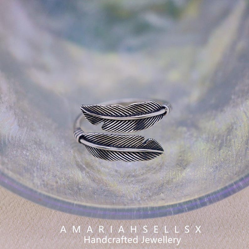 Puede incluir: Un anillo plateado con dise&ntilde;o de pluma. El anillo presenta dos motivos de plumas detallados, uno arriba y otro abajo, con l&iacute;neas intrincadas. El anillo se muestra sobre una superficie reflectante. El texto "AMARIAHSELLSX Handcrafted Jewellery" es visible.
