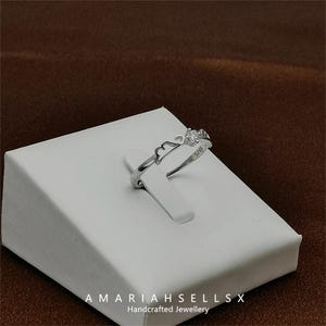 May include: A silver ring with a delicate design featuring a crown and the inscription "925". The ring is displayed on a white display stand with the text "AMARIAHSELLSX Handcrafted Jewellery".
