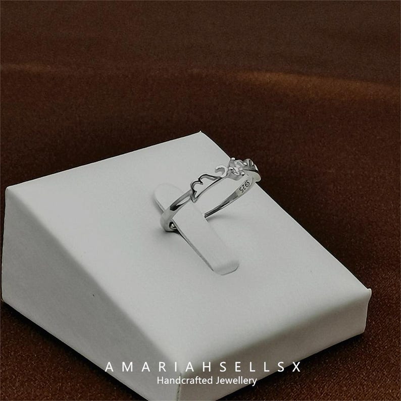 May include: A silver ring with a delicate design featuring a crown and the inscription "925". The ring is displayed on a white display stand with the text "AMARIAHSELLSX Handcrafted Jewellery".