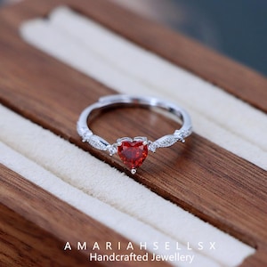 Op de afbeelding: Een zilveren ring met een rode hartvormige edelsteen en kleine heldere stenen op de band. De ring bevindt zich in een houten doos. AMARIAHSELLS Handgemaakte sieraden