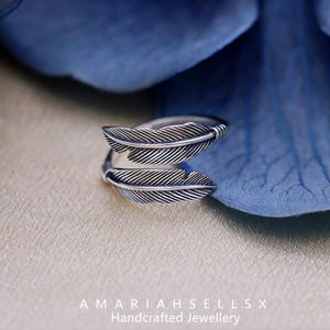 Puede incluir: Un anillo plateado con un dise&ntilde;o de doble pluma. Las plumas tienen l&iacute;neas detalladas, creando una apariencia texturizada. El anillo est&aacute; sobre un fondo suave y neutro con un acento floral azul. El texto "AMARIAHSELLSX Handcrafted Jewellery" es visible.