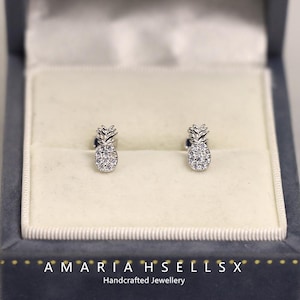 Puede incluir: Un par de pendientes de botón plateados con forma de piña, cada uno incrustado con pequeños cristales brillantes. Los pendientes se muestran dentro de una caja de joyería gris con el texto "AMARIAH SELLS X Handcrafted Jewellery".