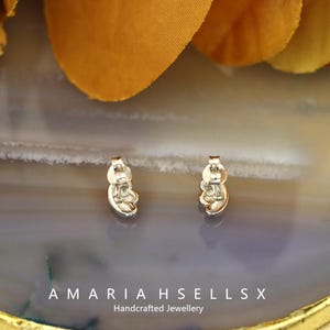 May include: A pair of silver stud earrings with a floral design. The earrings are set against a grey and white surface, with a blurred orange and yellow backdrop. The text "AMARIAH SELLS X Handcrafted Jewellery" is visible.