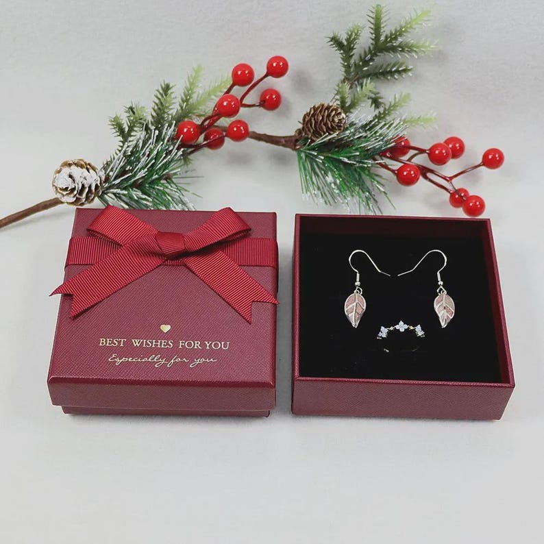 Puede incluir: Una caja de regalo granate con una cinta a juego y el texto "BEST WISHES FOR YOU, Especially for you." En el interior, se exhiben pendientes con forma de hoja y un anillo. La vegetaci&oacute;n con bayas rojas decora el fondo, creando una escena festiva.