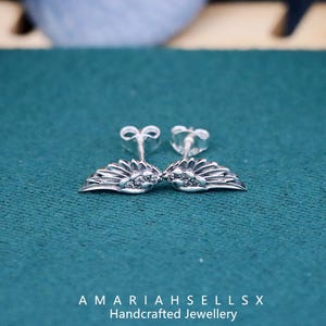 May include: A pair of silver earrings with a wing design and small clear stones. The earrings are on a green surface.